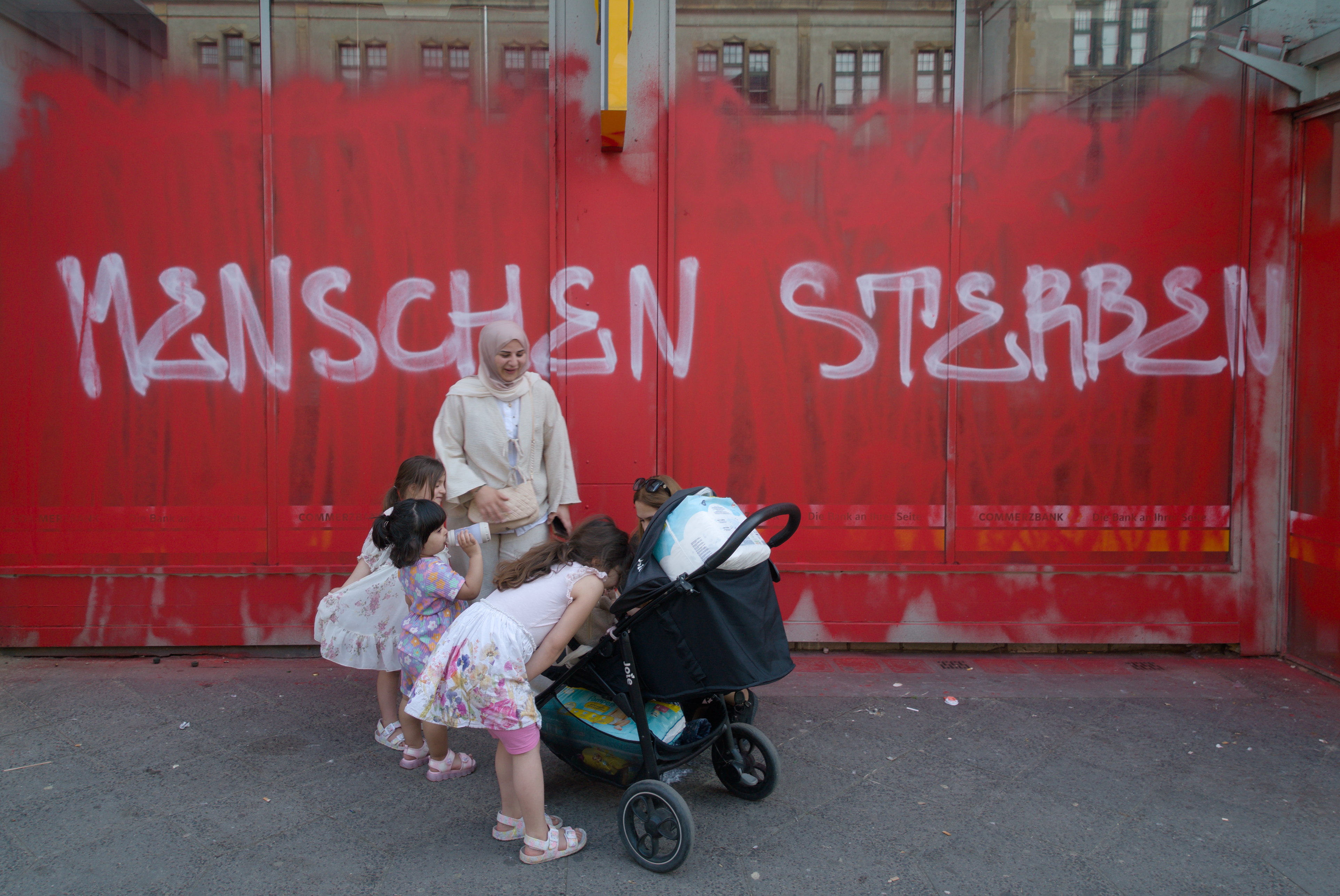 Madres e hijas entorno a un bebé. 'Menschen sterben' ha pintado alguien en la ventana de una sucursal bancaria. Serie 'el soporte' de Ibán Ojea. Berlín.