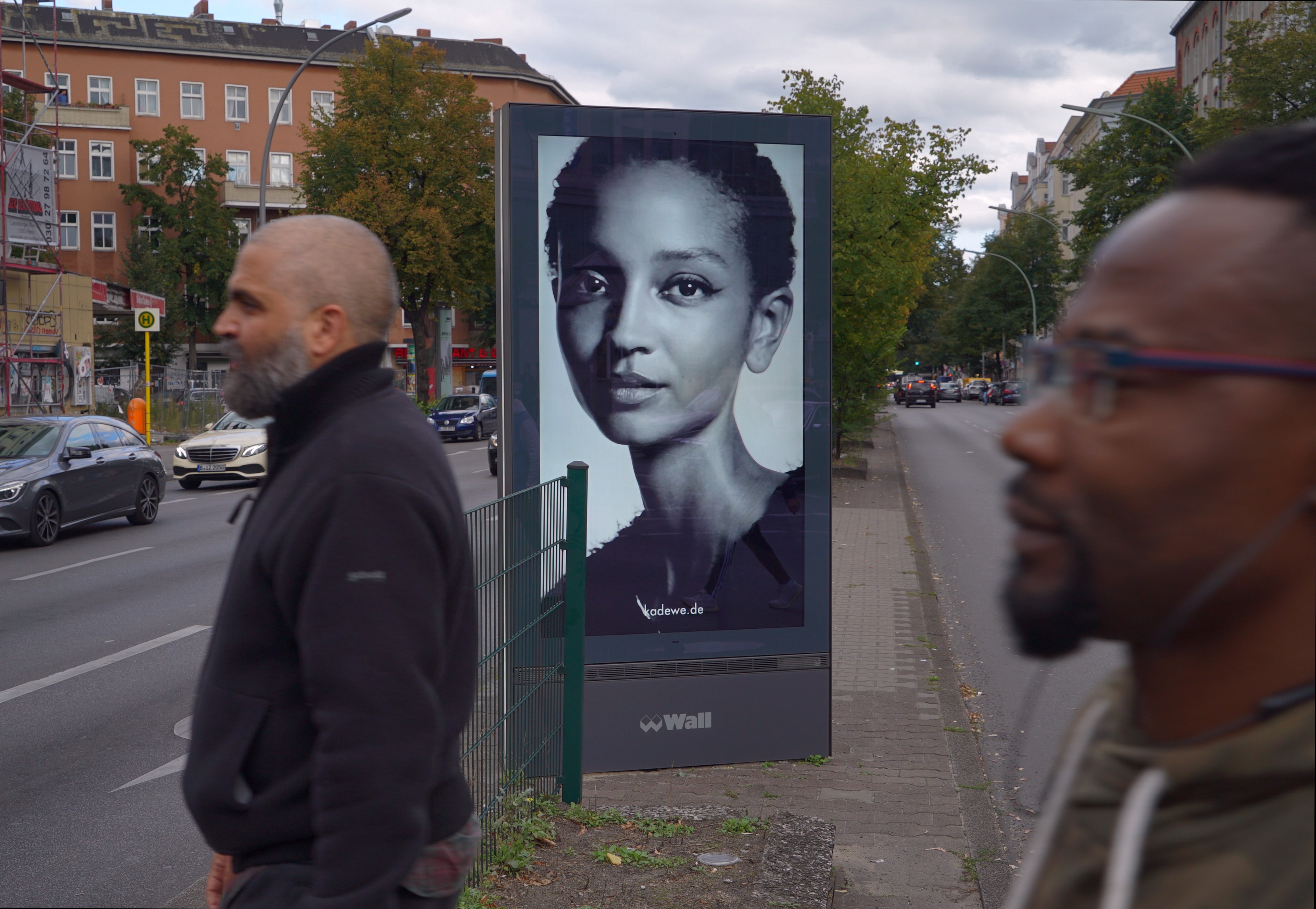 Dos personas pasan en primer término por delante de cámara. Detrás vemos una pantalla publicitaria en la que se ve el primer plano de una mujer que nos mira fijamente a nosotros. Serie 'el soporte' de Ibán Ojea.